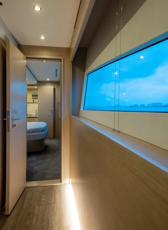 Interior view of a modern yacht's corridor, featuring a doorway and large window with a cloudy sky visible outside.